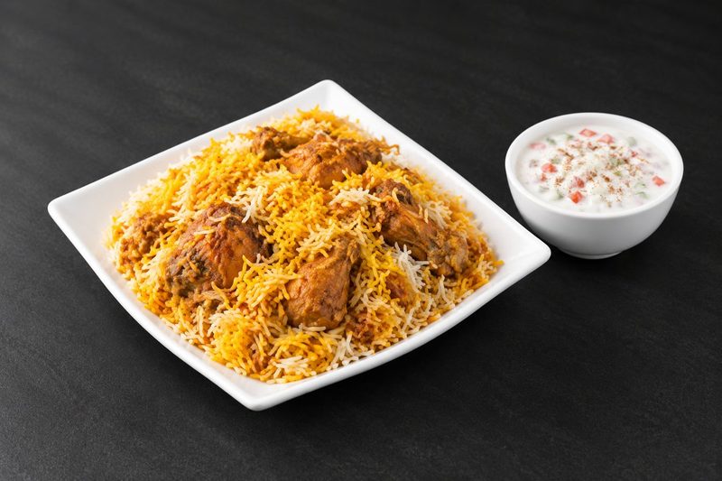Chicken biryani with saffron and white basmati rice on a square plate, with a bowl of raita alongside