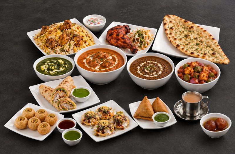 A spread of North Indian dishes on dark slate — chicken biryani with raita, tandoori chicken, naan, butter chicken, palak paneer, dal makhani, chilli paneer, samosas, pani puri, papdi chaat, masala chai and gulab jamun