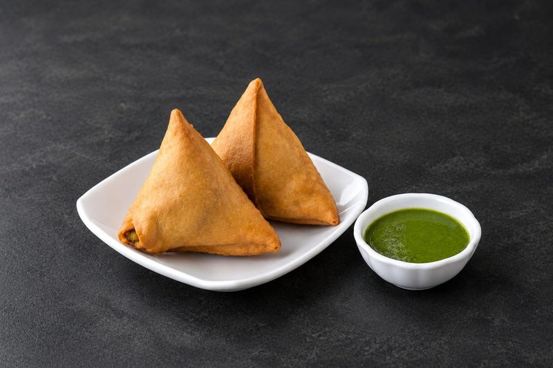 Two golden fried samosas on a white plate with a bowl of green mint chutney
